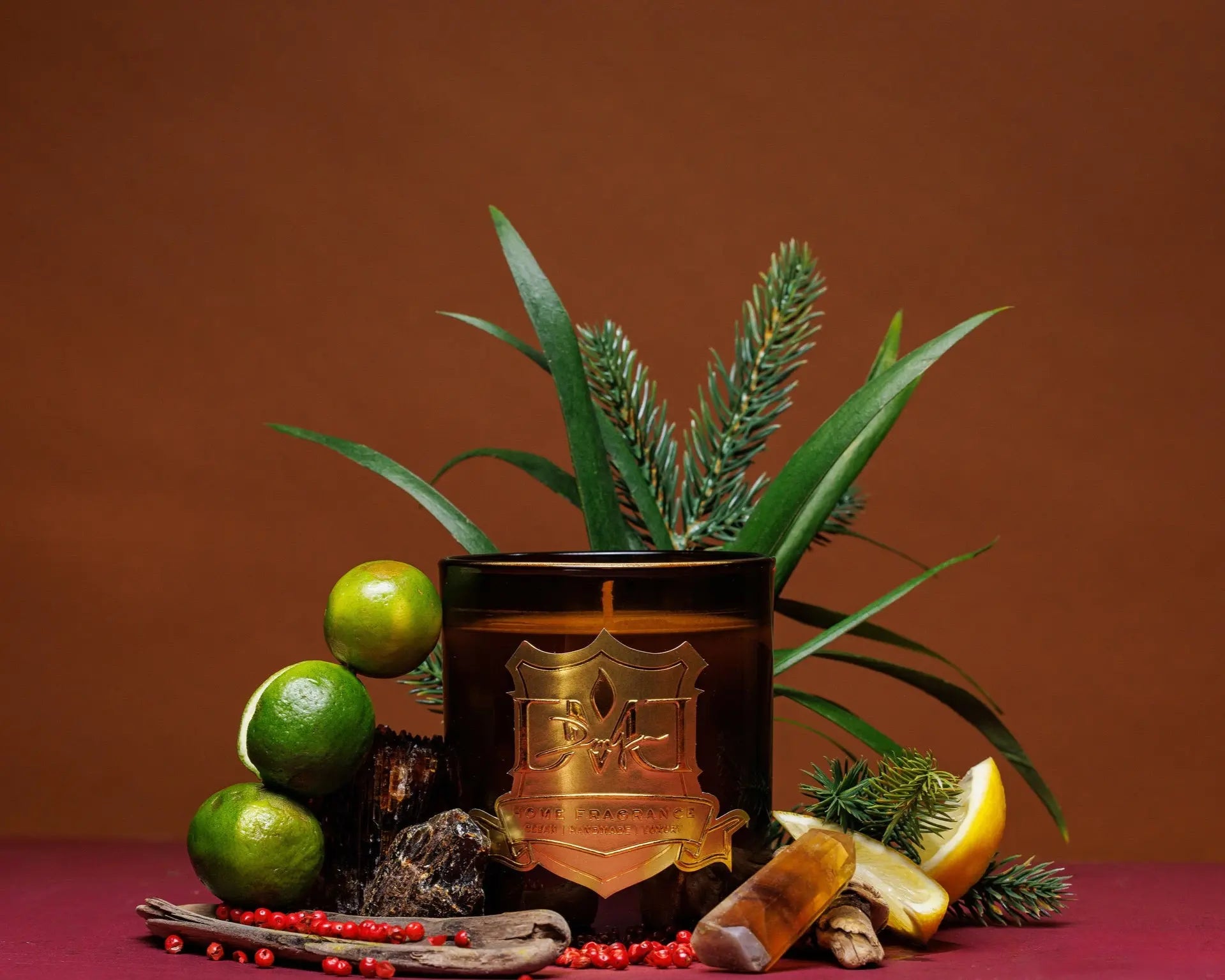 A scented candle with a brown tint, surrounded by decorative items including limes, a lemon, pine cones, and what appears to be a cinnamon stick, arranged on a flat surface. DMJ Home Fragrance candle. Luxury Clean Candle.