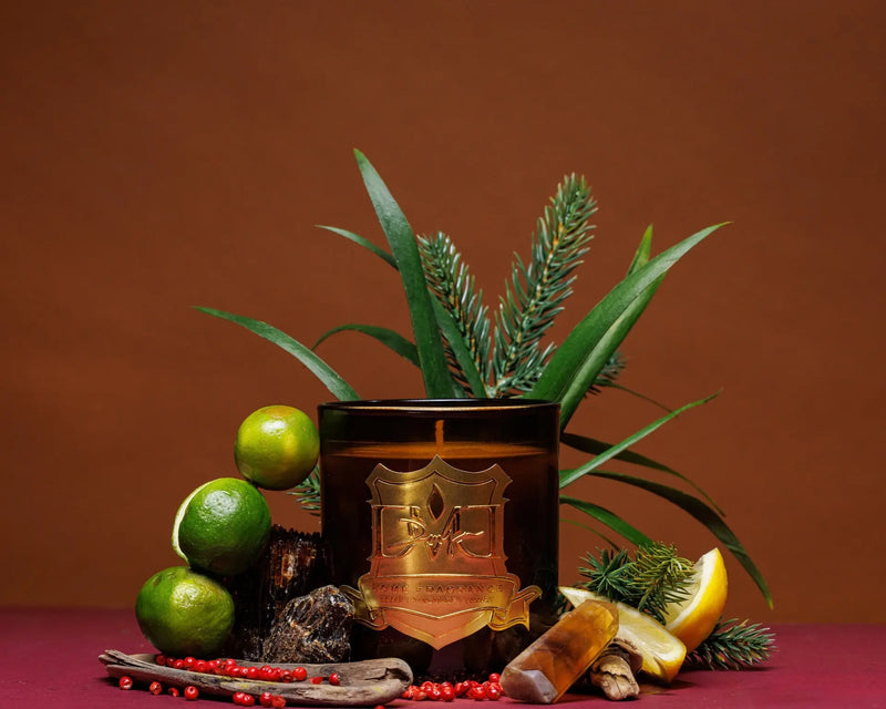 A scented candle with a brown tint, surrounded by decorative items including limes, a lemon, pine cones, and what appears to be a cinnamon stick, arranged on a flat surface. DMJ Home Fragrance candle. Luxury Clean Candle.