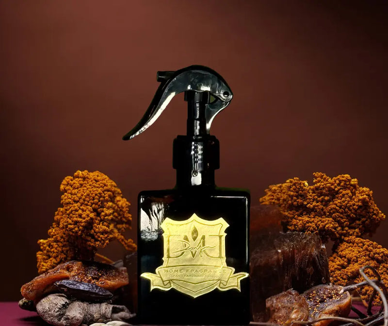 Two bottles of room spray with spray nozzles, one with a clear liquid and another with a black liquid, both featuring gold-colored labels. DMJ Home Fragrance Room Spray.