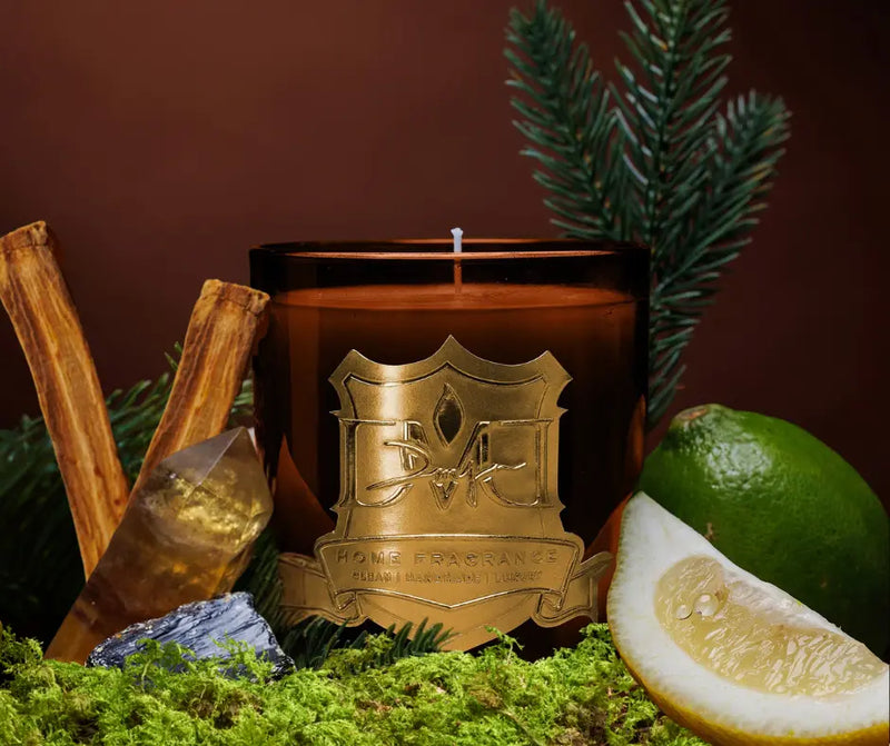 Candle with decorative emblem surrounded by cinnamon sticks, a lime, and lemon on a dark background
