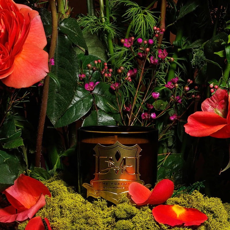 A candle with floral decorations, set in an ornate container, surrounded by various plants and flowers. DMJ Home Fragrance candle. Luxury Clean Candle.