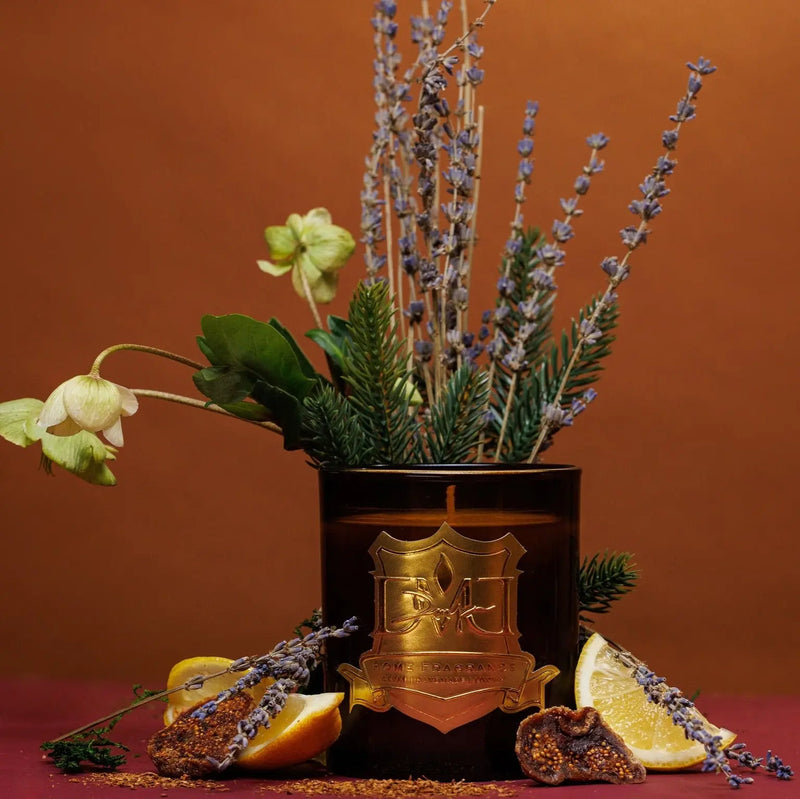 A candle with a bronze container, adorned with botanical decorations including lavender sprigs and lemon slices, set against a brown background. DMJ Home Fragrance candle. Luxury Clean Candle.