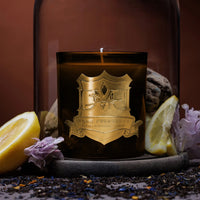 A scented candle with a bergamot fragrance, placed in a clear glass jar with a gold lid, surrounded by lemon slices and violet leaves on a dark surface. DMJ Home Fragrance candle. Luxury Clean Candle.
