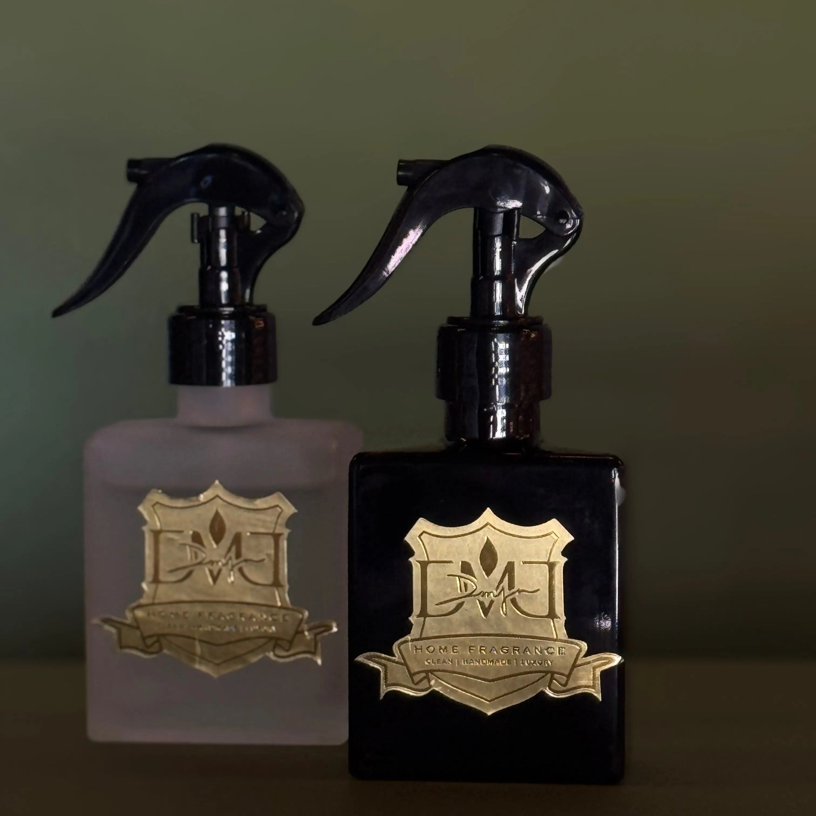 Two bottles of room spray with spray nozzles, one with a clear liquid and another with a black liquid, both featuring gold-colored labels. DMJ Home Fragrance Room Spray.