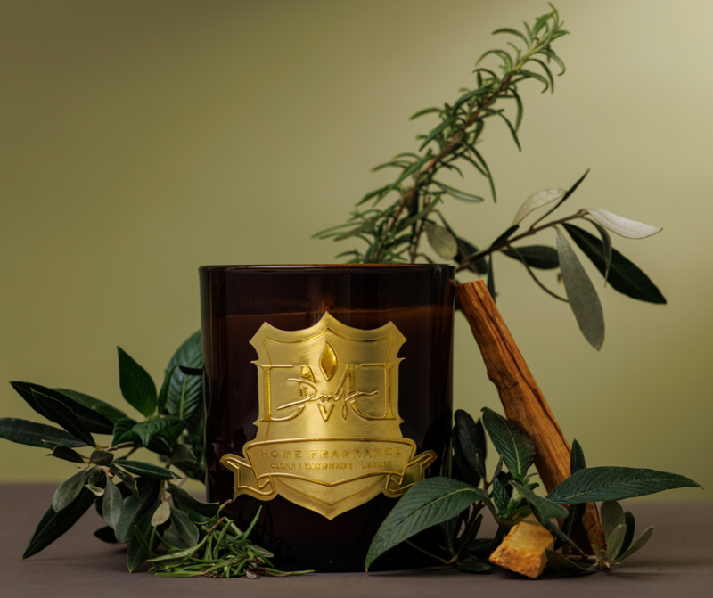 Dark brown candle with gold emblem surrounded by green leaves and a cinnamon stick on a beige background