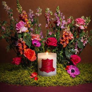 DMJ Home Fragrance luxury candle lit and surrounded by vibrant roses and wild florals on moss, styled for a high-end product shoot showcasing clean coconut wax craftsmanship and refined home fragrance design.