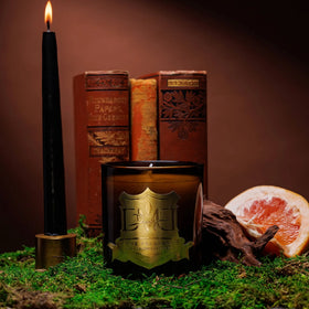 Hemingway candle by DMJ Home Fragrance in amber glass with gold crest beside antique books, a black taper and grapefruit
