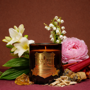 Luxury Kyoto candle by DMJ Home Fragrance in an amber glass vessel with gold label, styled with freesia, lily of the valley, pink peony, rhubarb stalks, blonde wood shavings, and raw amber stones on a warm brown background, showcasing clean luxury candle fragrance notes and artisanal home fragrance styling.