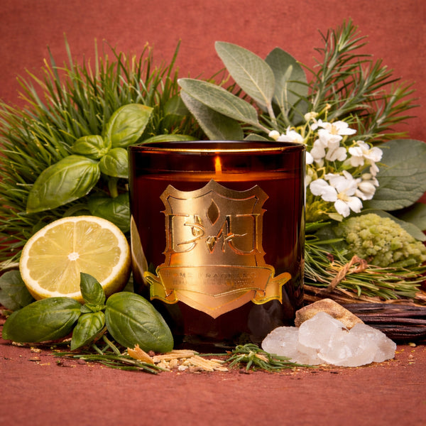 Candle with decorative label surrounded by greenery, lemon, and white flowers on a red background