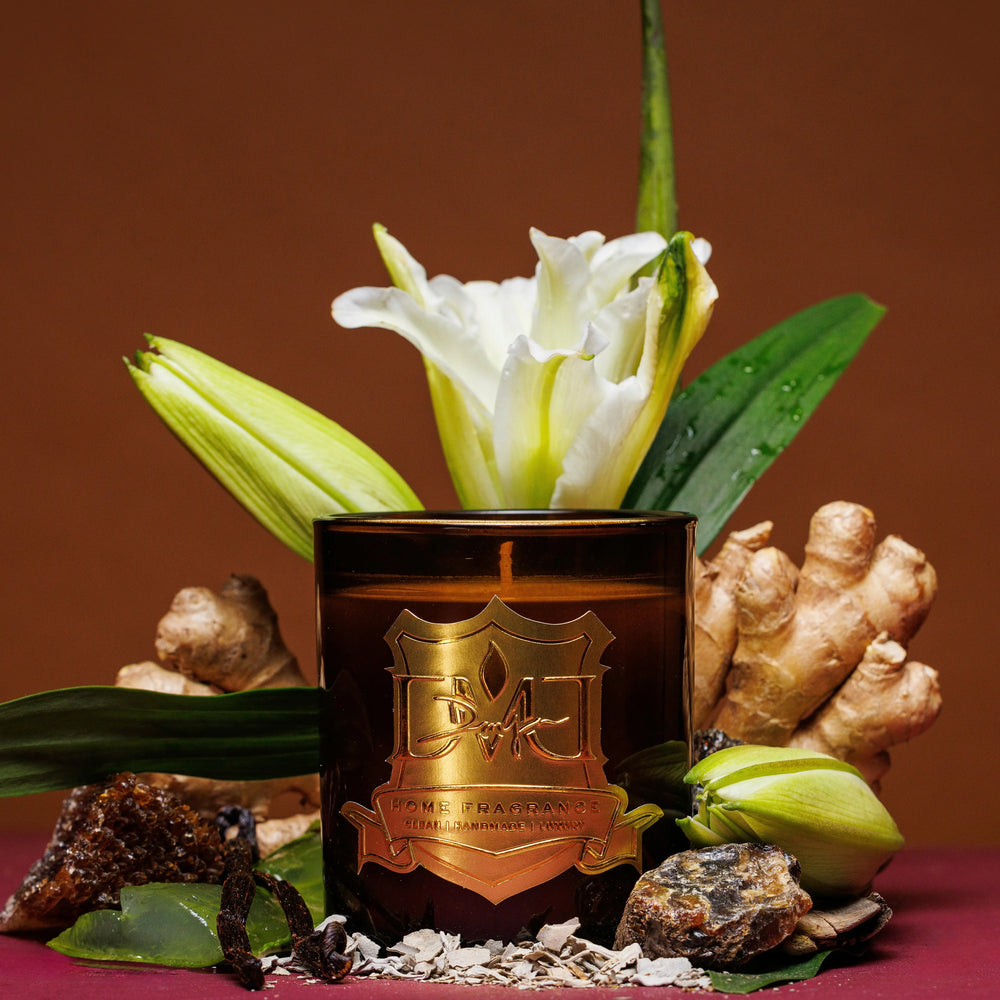 A scented candle with a floral decoration on top, surrounded by greenery and a ginger root, on a dark background. DMJ Home Fragrance candle. Luxury Clean Candle.