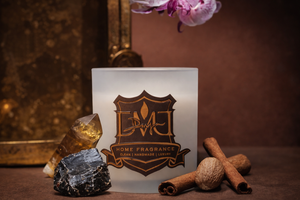 Luxury DMJ Home Fragrance candle in a frosted white glass vessel with gold logo, styled with cinnamon sticks, nutmeg, and crystal stones, photographed in warm moody lighting for Mood by DMJ in Lafayette Louisiana, showcasing clean handmade luxury home fragrance and artisan candle design.