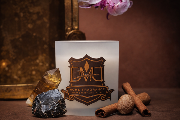Luxury DMJ Home Fragrance candle in a frosted white glass vessel with gold logo, styled with cinnamon sticks, nutmeg, and crystal stones, photographed in warm moody lighting for Mood by DMJ in Lafayette Louisiana, showcasing clean handmade luxury home fragrance and artisan candle design.