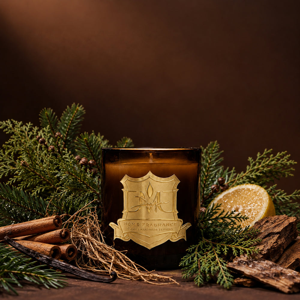 Winter Wood Candle by DMJ Home Fragrance in dark amber glass with a gold crest label, styled with cypress and fir branches, cinnamon sticks, vanilla pod, vetiver root, halved citron and cedar bark on a warm brown backdrop