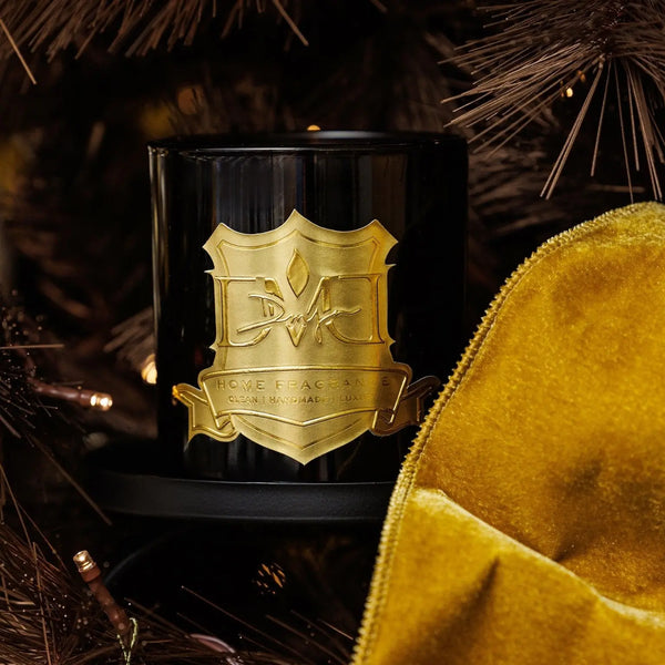 Winter Woods holiday candle by DMJ Home Fragrance in black glass with gold crest among pine branches and gold velvet