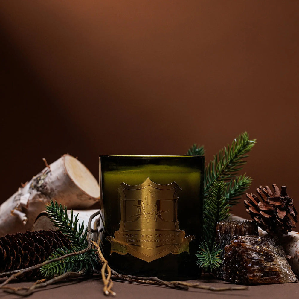 Woods pine and fir balsam candle by DMJ Home Fragrance in green glass with birch logs, pine sprigs, and pinecones